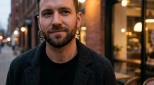 Close-up of a stylish man in his 30s, wearing a dark jacket and modern hoop earrings, with a confident expression in a blurred urban setting.