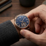 Remonter une montre automatique : la méthode simple et sûre 3 Close-up of a well-groomed hand winding a rose gold automatic watch with a dark blue dial, Roman numerals, and visible movement. Blurred wood background.