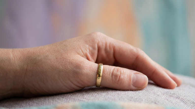 Close-up of a relaxed female hand with a gold thumb ring, against a soft, blurred background of muted lavender, peach, and teal.