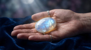 A hand gently cradles a polished moonstone displaying ethereal blue, white, and silver shimmers, glowing warmly against dark velvet.