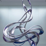 Couleur argent : signification, symbolique et élégance 3 Elegant silver abstract sculpture with flowing, polished ribbons reflecting soft light and subtle blue/purple hues, set in a minimalist space.