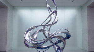 Elegant silver abstract sculpture with flowing, polished ribbons reflecting soft light and subtle blue/purple hues, set in a minimalist space.