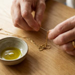Démêler une chaîne : l'astuce miracle à l'huile d'olive 3 Close-up of gentle hands meticulously untangling a delicate gold chain with a needle, next to a ceramic bowl of olive oil on a wooden table.