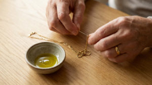 Close-up of gentle hands meticulously untangling a delicate gold chain with a needle, next to a ceramic bowl of olive oil on a wooden table.