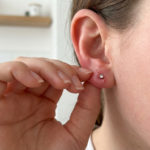 Infection boucle oreille : comment sauver son piercing ? 3 Oreille percée avec signes d'infection débutante. Une main aux doigts soignés touche la zone, fond flou et lumineux.