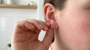 Infection boucle oreille : comment sauver son piercing ? 2 Oreille percée avec signes d'infection débutante. Une main aux doigts soignés touche la zone, fond flou et lumineux.