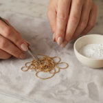 Démêler un collier : techniques pour sauver vos bijoux 3 Well-manicured hands delicately detangling a tangled gold chain with tools on a soft gray surface, next to talcum powder and oil.