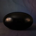 Onyx noire : signification et pouvoirs de protection 1 A highly polished, deep black onyx stone, oval-shaped, reflecting soft cinematic light. It rests on a textured dark surface against a muted indigo and plum background.