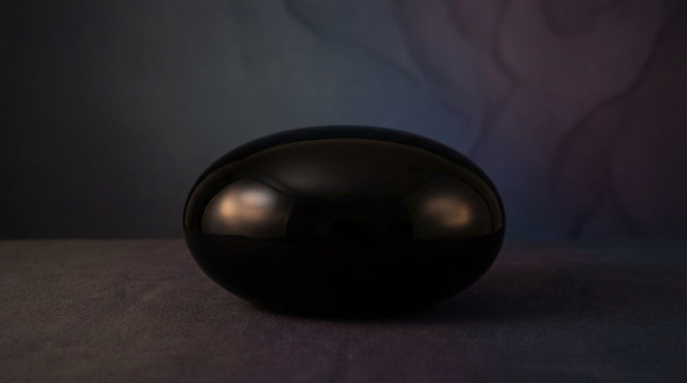 A highly polished, deep black onyx stone, oval-shaped, reflecting soft cinematic light. It rests on a textured dark surface against a muted indigo and plum background.