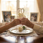 Noces de platine : célébrer 70 ans d'amour éternel 7 Elderly couple holding hands across a table, celebrating their 70th platinum wedding anniversary. Rings, champagne, and '70' centerpiece visible.
