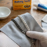 Nettoyer une chaîne en argent : secrets pour briller 3 Gloved hands polish a silver chain with a cloth. One half is dark and tarnished, the other sparkling clean. Baking soda and toothpaste are visible.