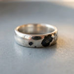 Votre bague en argent noircit ? La vérité sur ce phénomène 4 A silver ring displaying shiny areas contrasted by distinct, irregular matte black tarnish patches, on a blurred grey background.