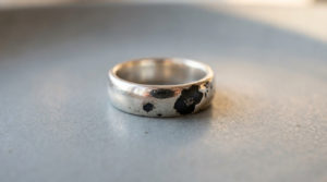 A silver ring displaying shiny areas contrasted by distinct, irregular matte black tarnish patches, on a blurred grey background.