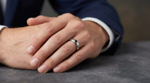 Close-up of a well-groomed male hand with a sophisticated silver ring and black gemstone on the ring finger. Suit cuff visible.