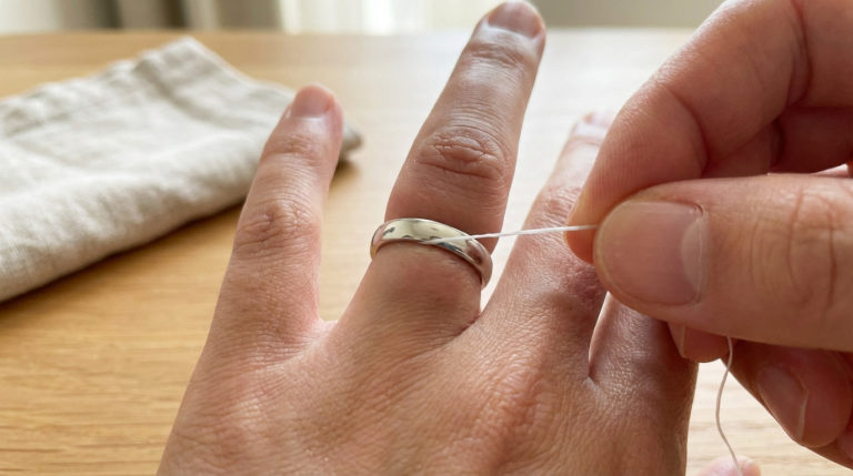 A close-up shows a hand with a tight silver ring on a swollen finger, as a white thread is carefully inserted to aid removal.