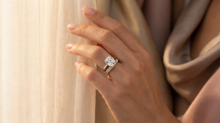 Close-up of an elegant hand wearing a sparkling cushion-cut diamond engagement ring and a pavé wedding band, glowing in soft, warm light against a blurred neutral background.